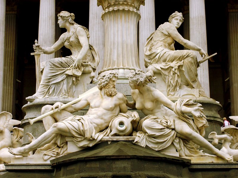 OLYMPUS DIGITAL CAMERA Fotografia em close das esculturas de mármore na base da Fonte de Pallas Athena, em Viena. Em primeiro plano, duas figuras reclinadas representam rios; acima delas, duas estátuas sentadas simbolizam o Poder Executivo e o Legislativo, com colunas clássicas ao fundo. Atena é uma personagem da SAga Percy jackson
